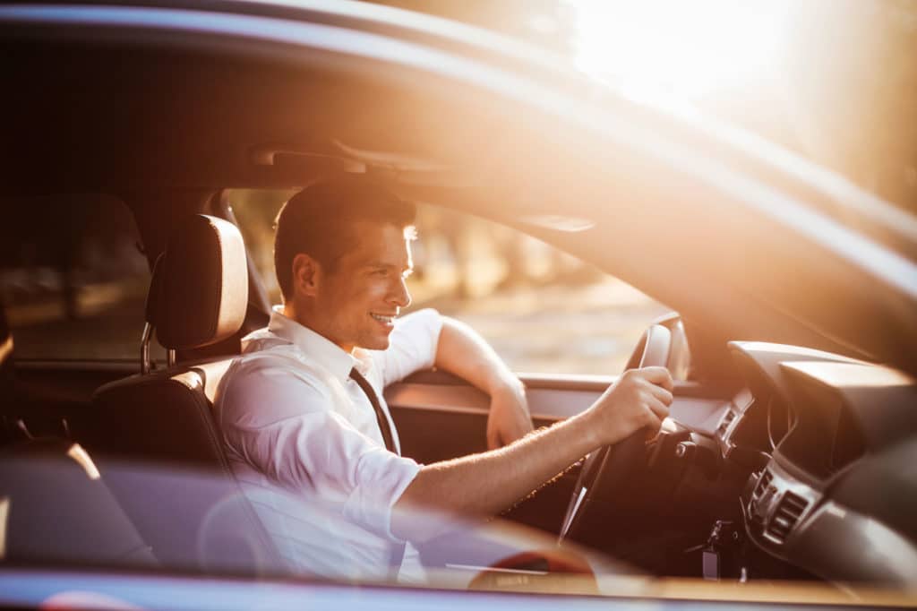 Man sitting at the wheel of a car with his right hand on the steering wheel and left elbow hanging out the window