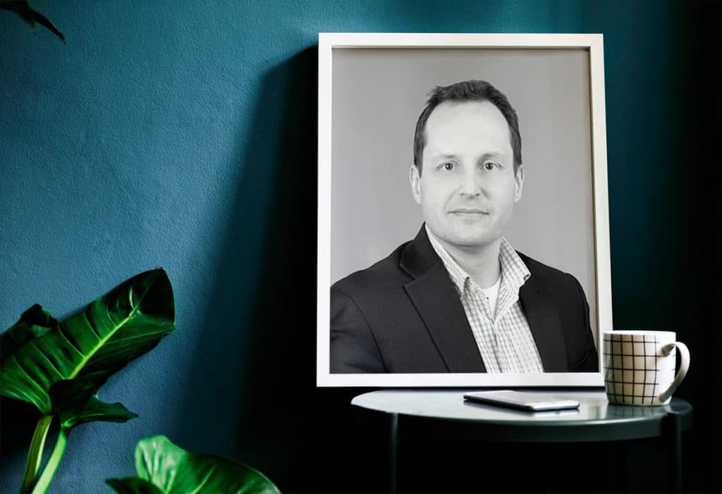 Picture of a man on a table with a mug and a phone in front of it and a plant in the corner