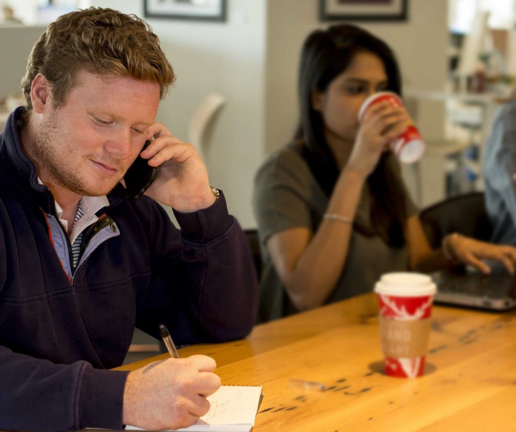 Man talking on a cell phone at a table with a woman drinking from a red coffee cup in background