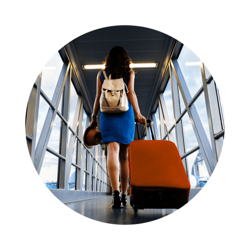Back view of a woman walking down a hallway pulling a wheeled suitcase and carrying a hat and a backpack